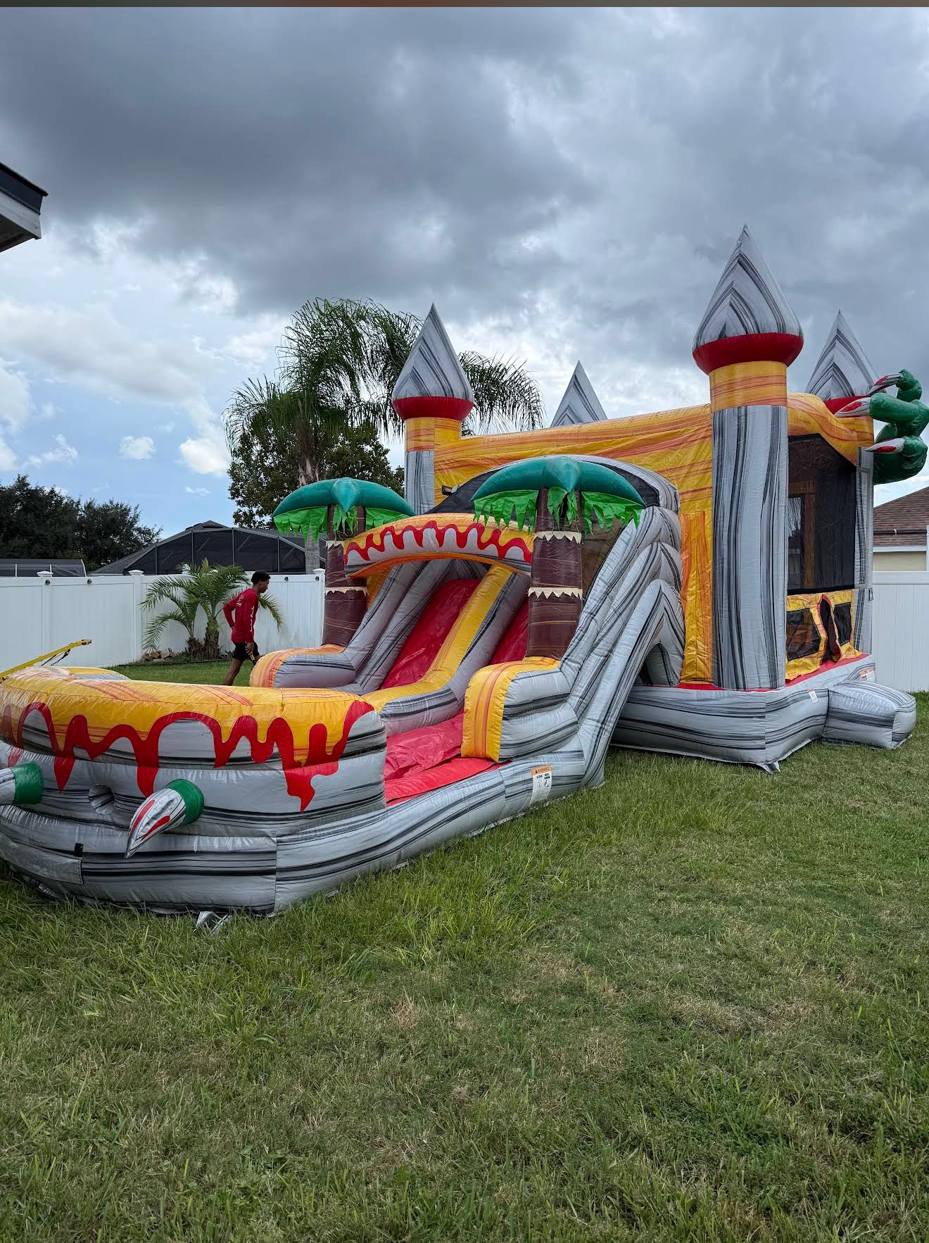 Dino bounce house and slide with gray towers, red and yellow lanes and green dinosaur